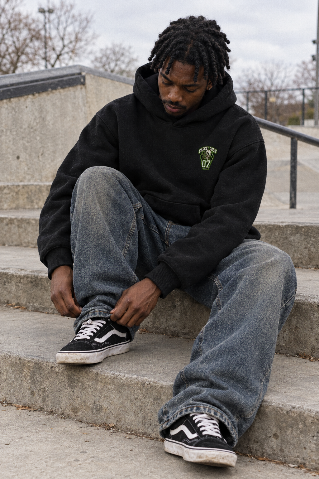 A man sitting on concrete steps wearing a black hoodie with a green chest graphic, loose blue jeans, and black Vans sneakers.