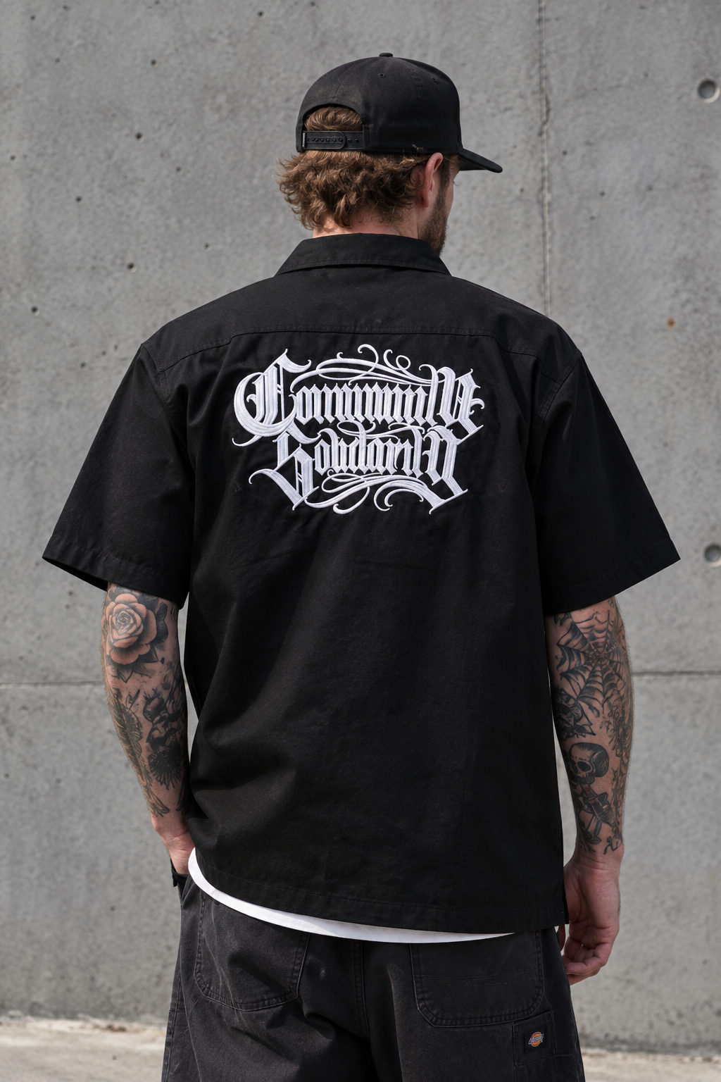 A man viewed from behind wearing a black short-sleeve button-up shirt with a large white gothic-style graphic reading 'Communauté Bordelaise' across the back, paired with a black cap and dark pants, standing against a concrete wall.