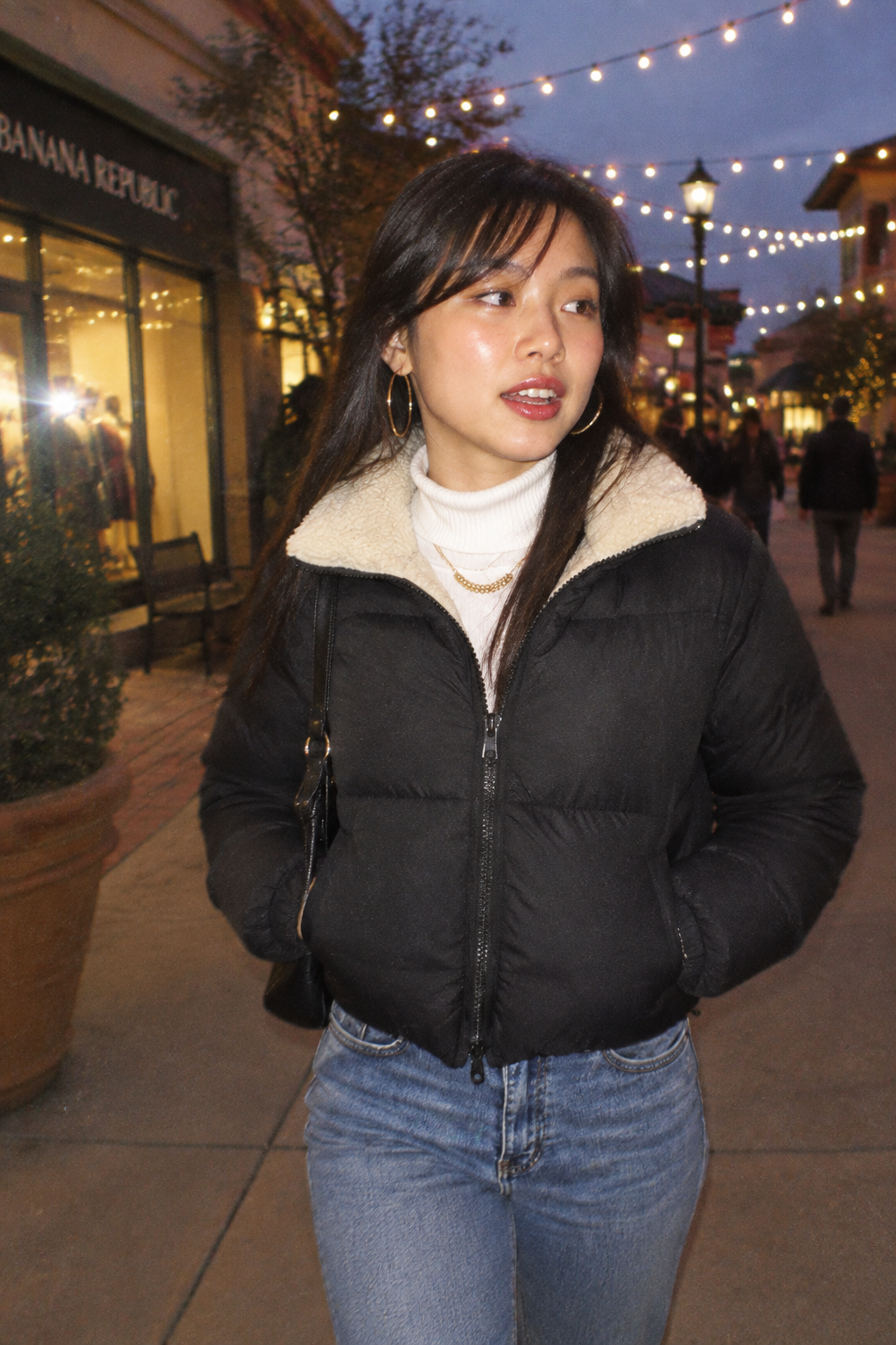 A woman wearing a black puffer jacket with a cream sherpa collar, a white turtleneck, and blue jeans, standing outdoors on a pedestrian shopping area at dusk.
