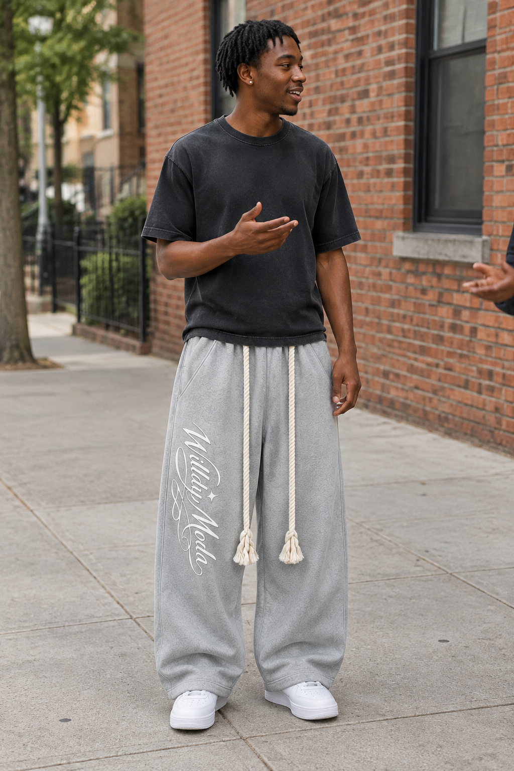 A man standing on a city sidewalk wearing a black t-shirt, light gray sweatpants with white drawstrings and script text, and white sneakers, with a brick building in the background.