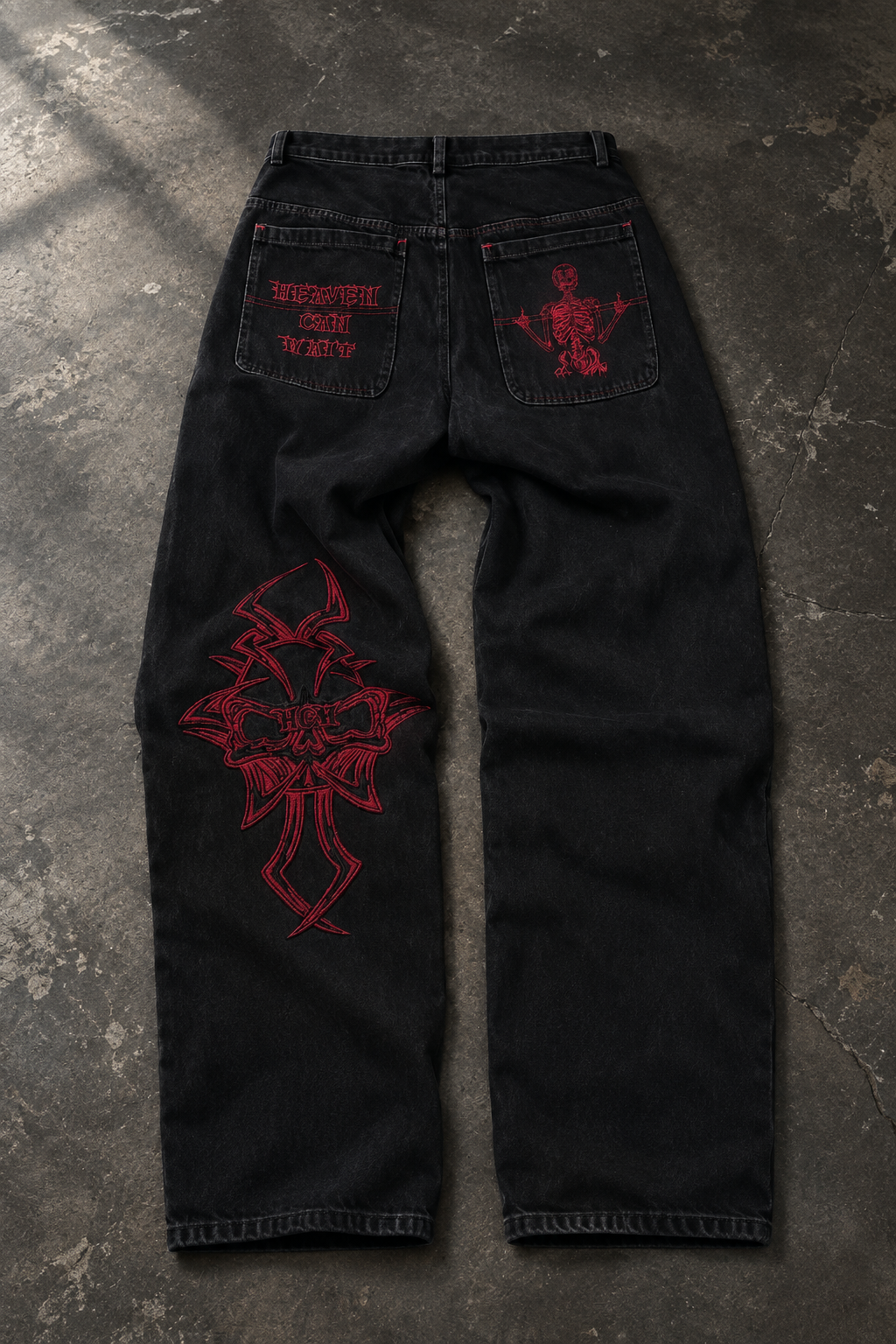 Black denim jeans laid flat on a textured gray concrete surface, featuring red embroidered graphics including a large cross motif on the right leg and red back pocket embroidery.