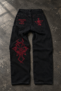 Black denim jeans laid flat on a textured gray concrete surface, featuring red embroidered graphics including a large cross motif on the right leg and red back pocket embroidery.