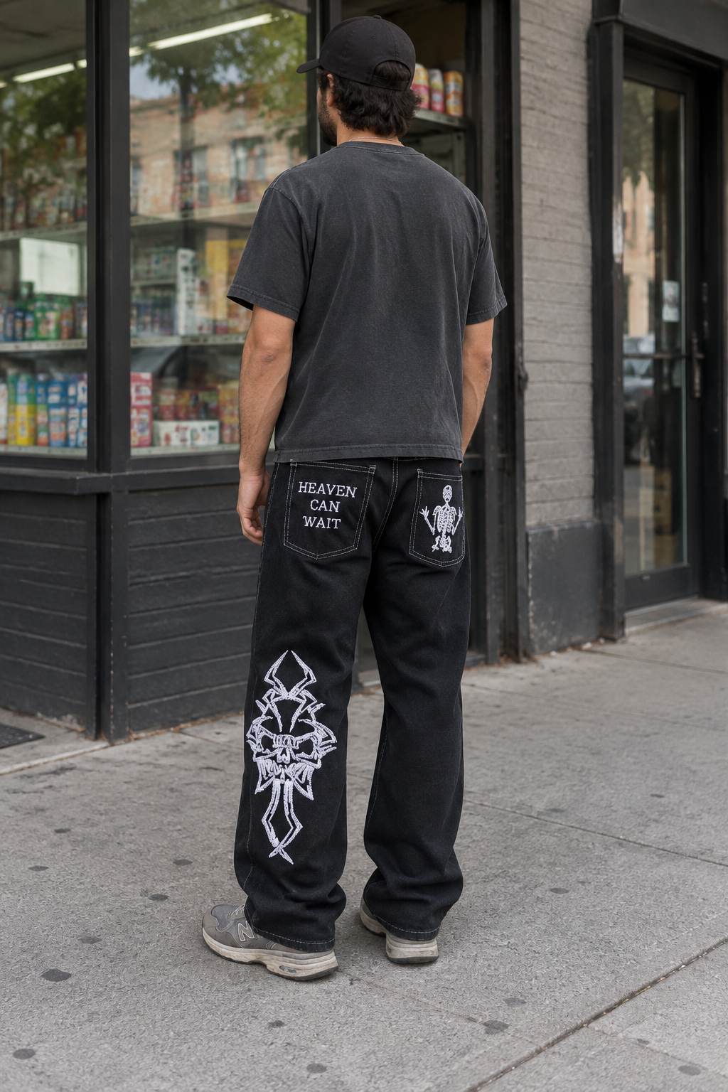 A man viewed from behind wearing black graphic sweatpants with white skeleton and text designs on the back pockets, a black t-shirt, and a black cap, standing on a city sidewalk in front of a store.