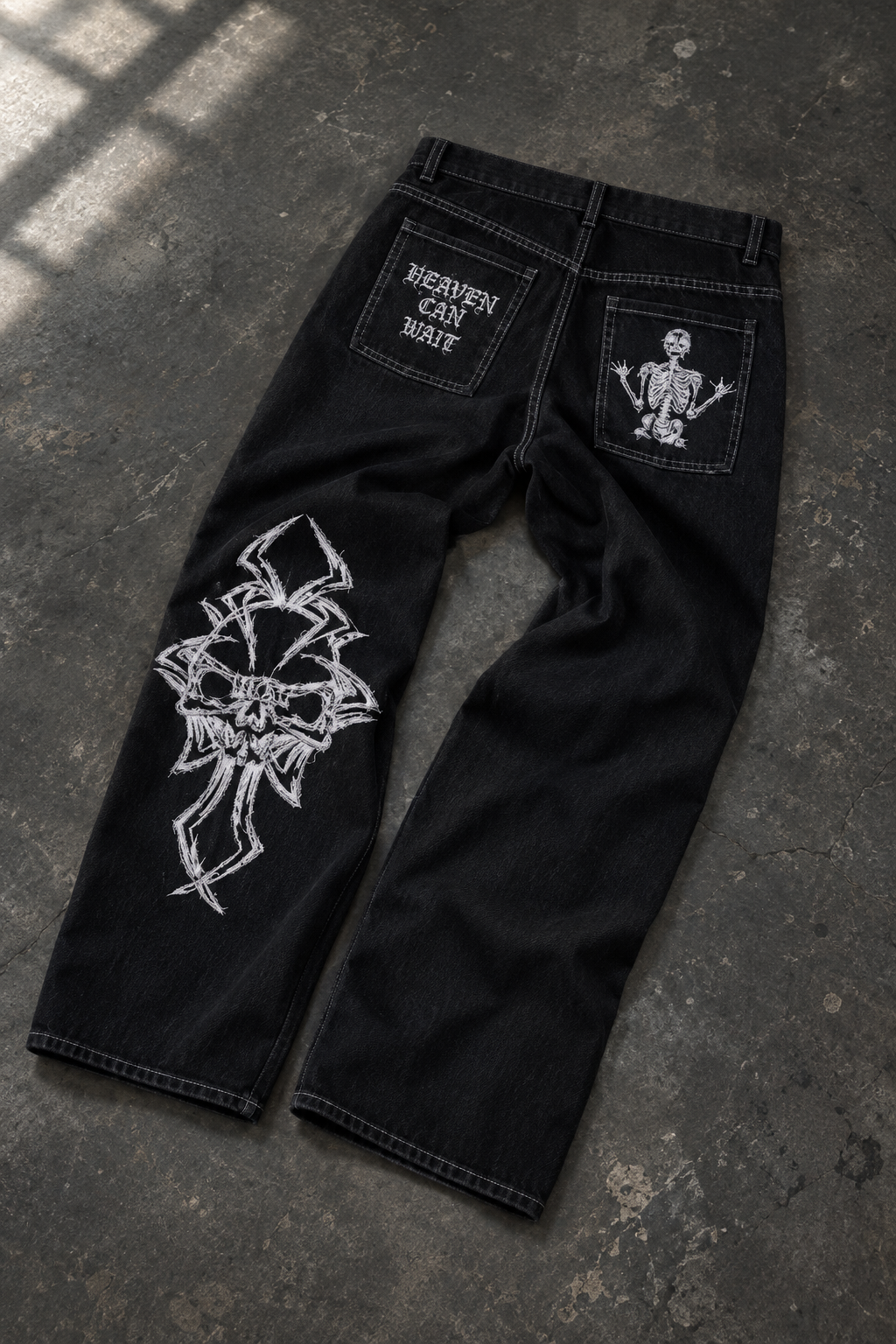 Black denim jeans laid flat on a dark textured floor, featuring a large white graphic print on the right leg, a small white skeleton patch on the back right pocket, and a white text patch on the back left pocket that reads 'HEAVEN CAN ONLY BE TENDED BY THE LIVING'.