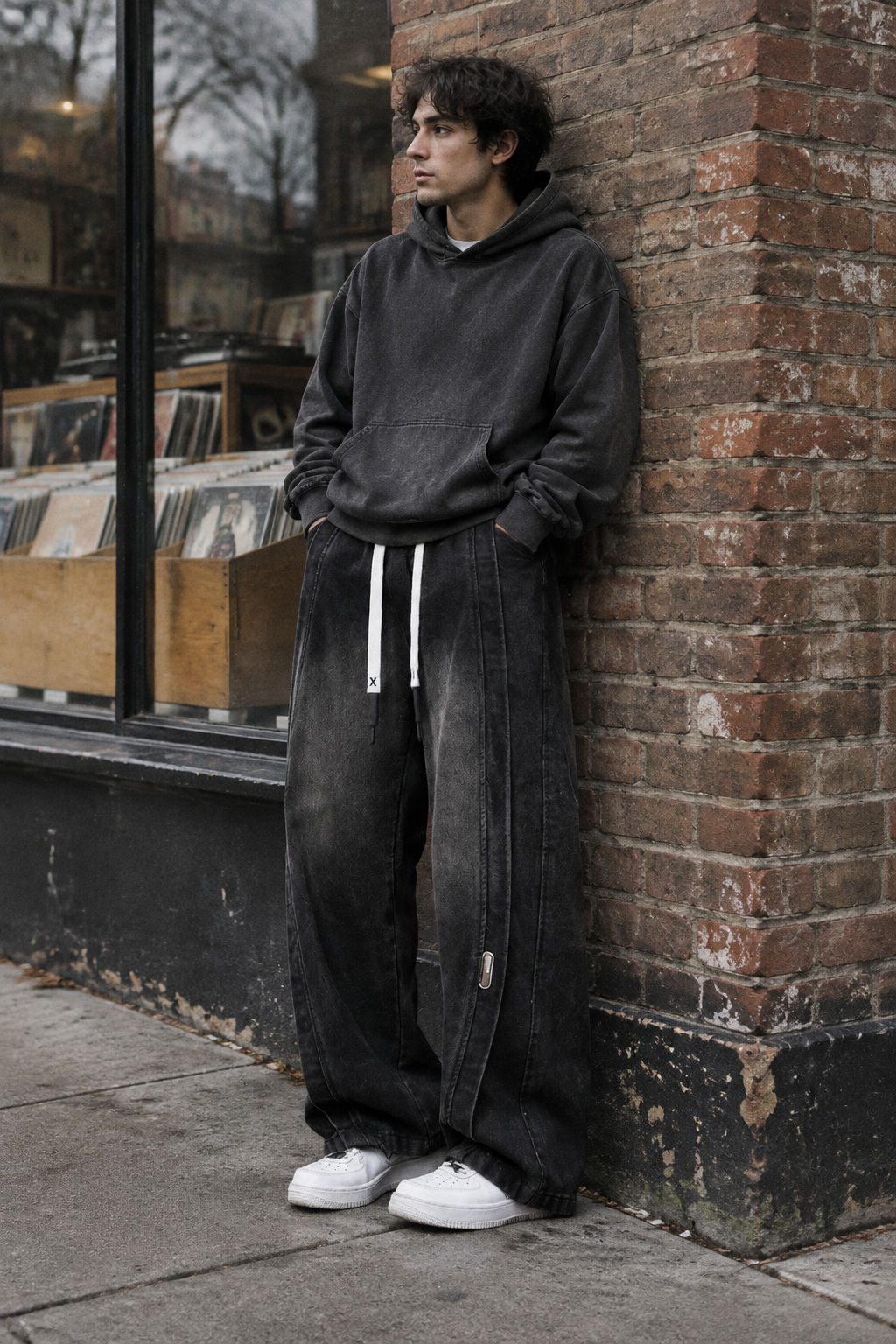 A man wearing a washed black hoodie and wide-leg black denim sweatpants with white drawstrings, leaning against a brick wall on a city sidewalk.