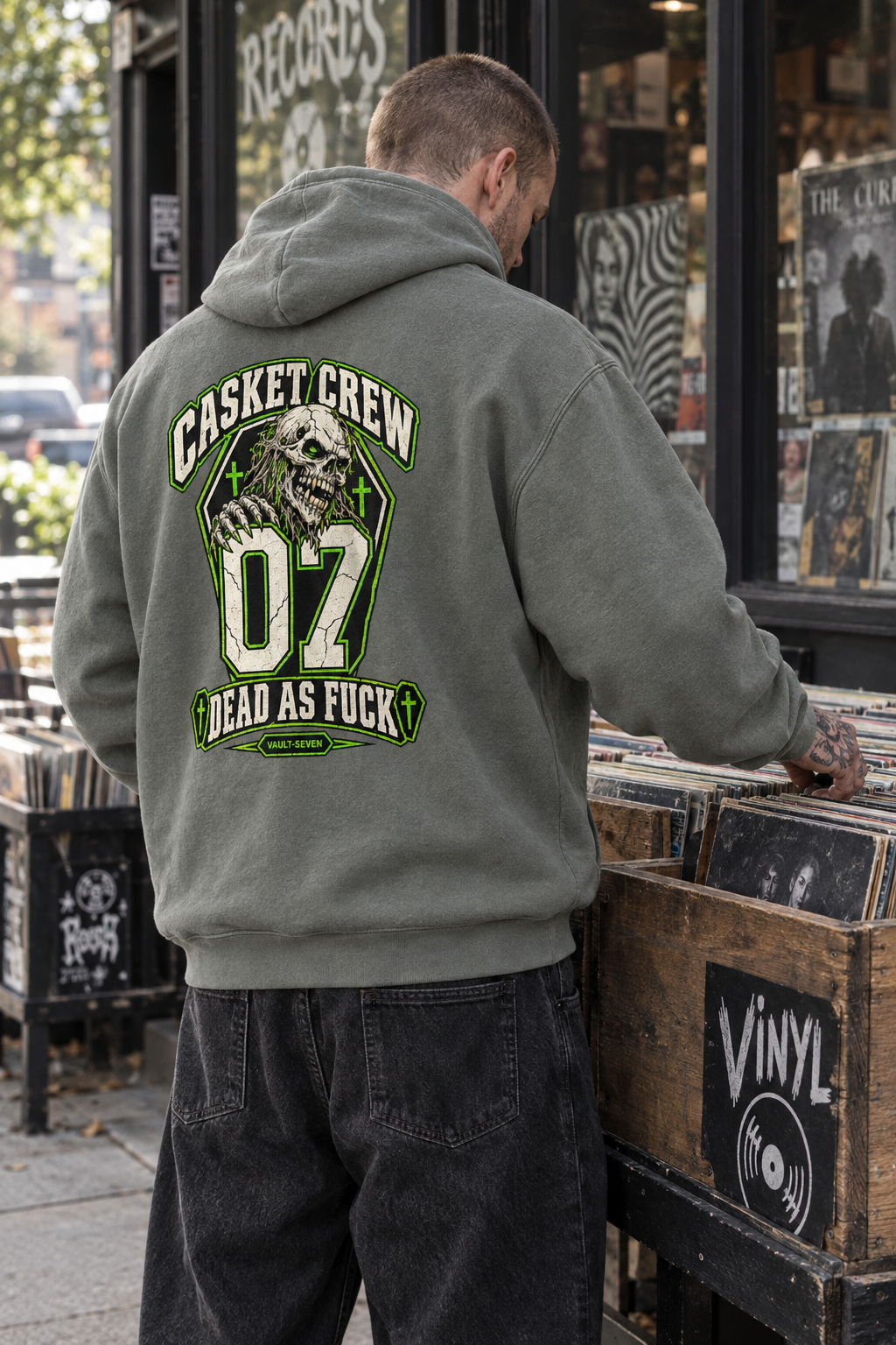 A man viewed from behind wearing an olive green hoodie with a large graphic on the back that reads 'CASKET CREW 07 DEAD AS FUCK' with a skull and green accents, standing in front of a vinyl record store window.