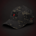 A dark camouflage baseball cap with 'VAULT SEVEN' embroidered on the front in black and red, set against a dark brown background.