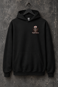 A black pullover hoodie hanging on a black hanger against a textured dark gray wall. The hoodie features a small chest graphic of a skull wearing a hard hat with a red '7' and the text 'Vault Seven' printed below it.