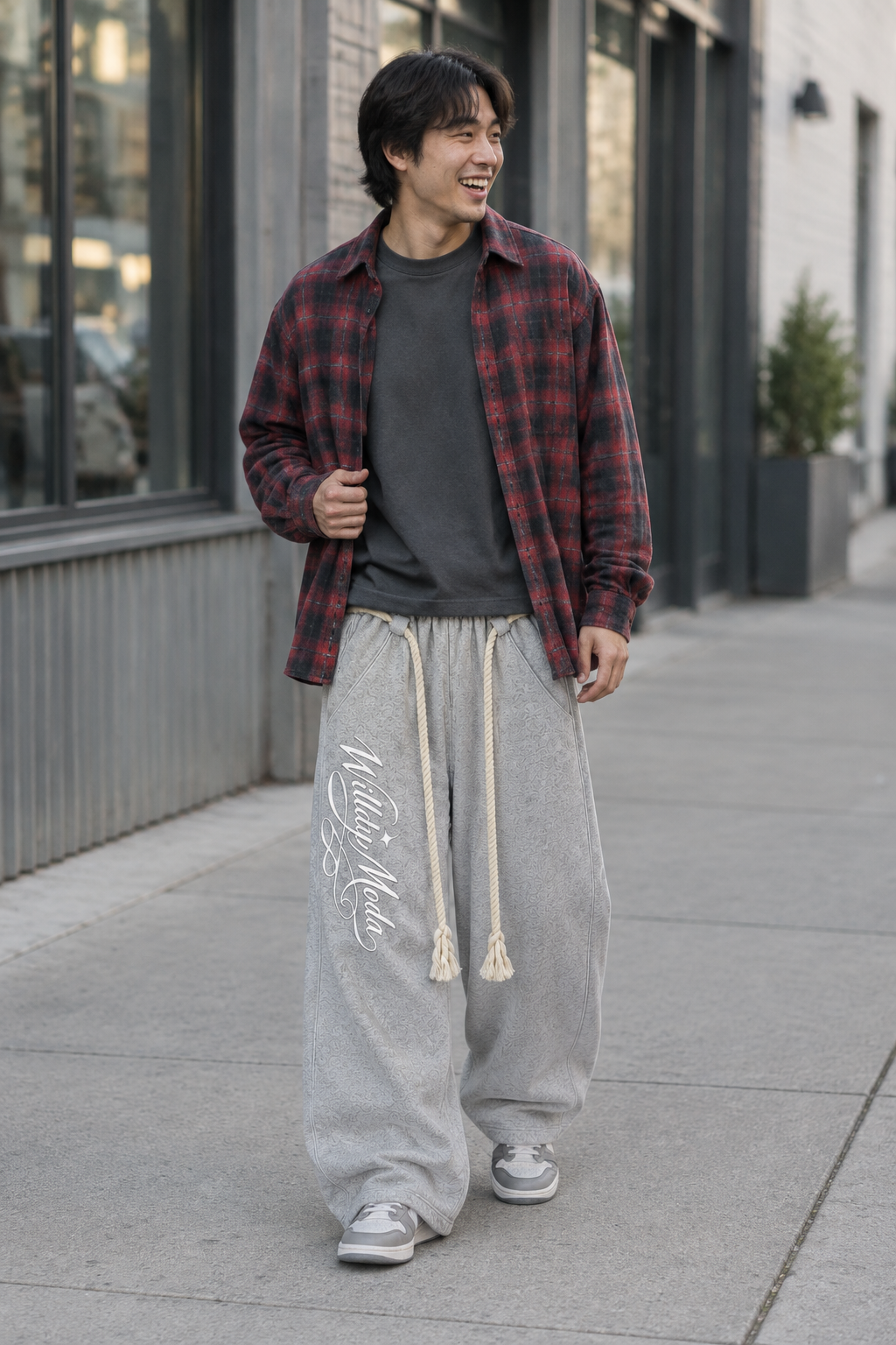 A man walking on a city sidewalk wearing a red and black plaid flannel shirt over a dark gray t-shirt, paired with baggy light gray sweatpants and white sneakers.