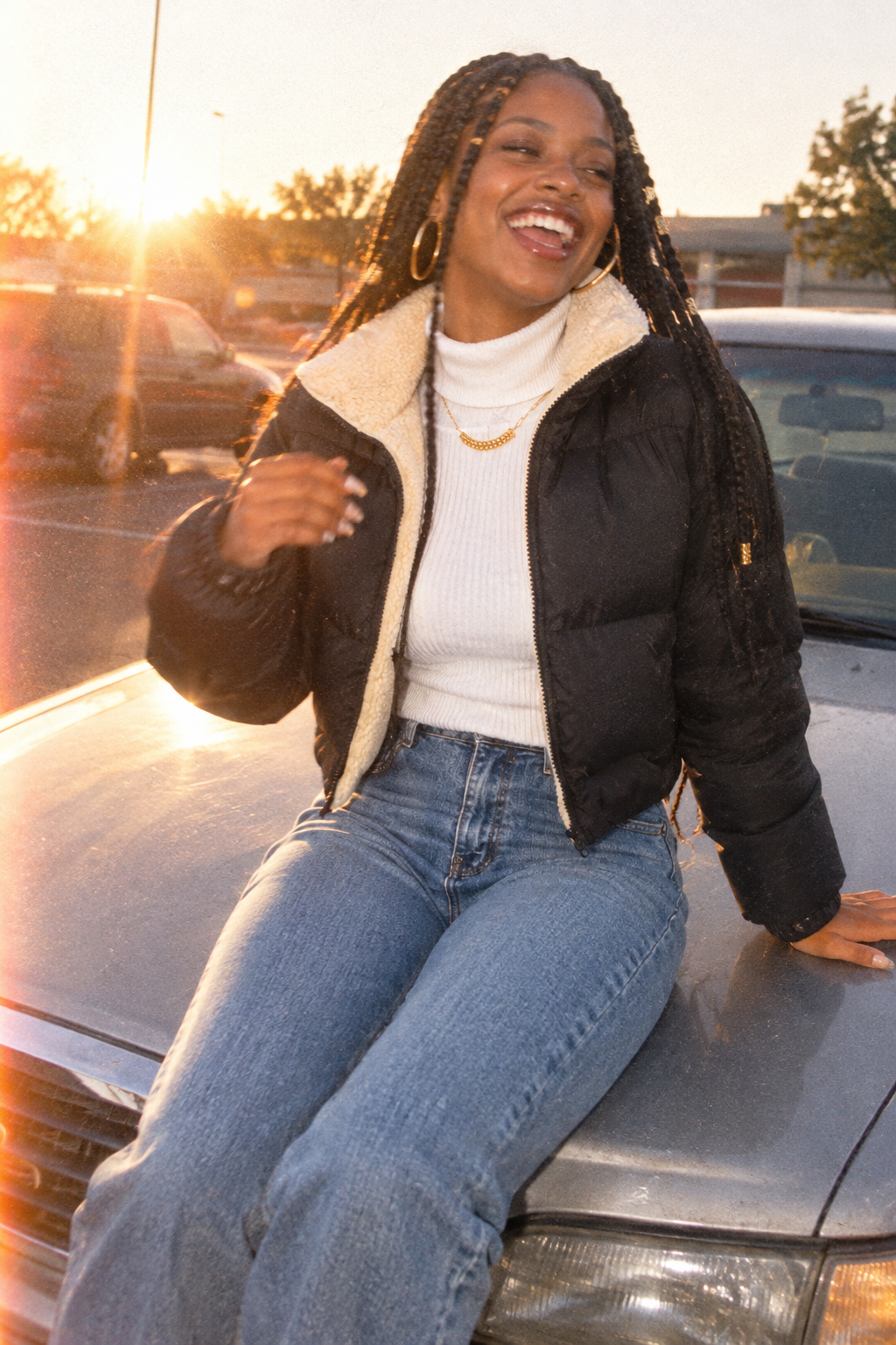 A woman smiling while sitting on the hood of a car in a parking lot at sunset, wearing a black puffer jacket with a cream sherpa lining, a white ribbed turtleneck, and blue jeans.