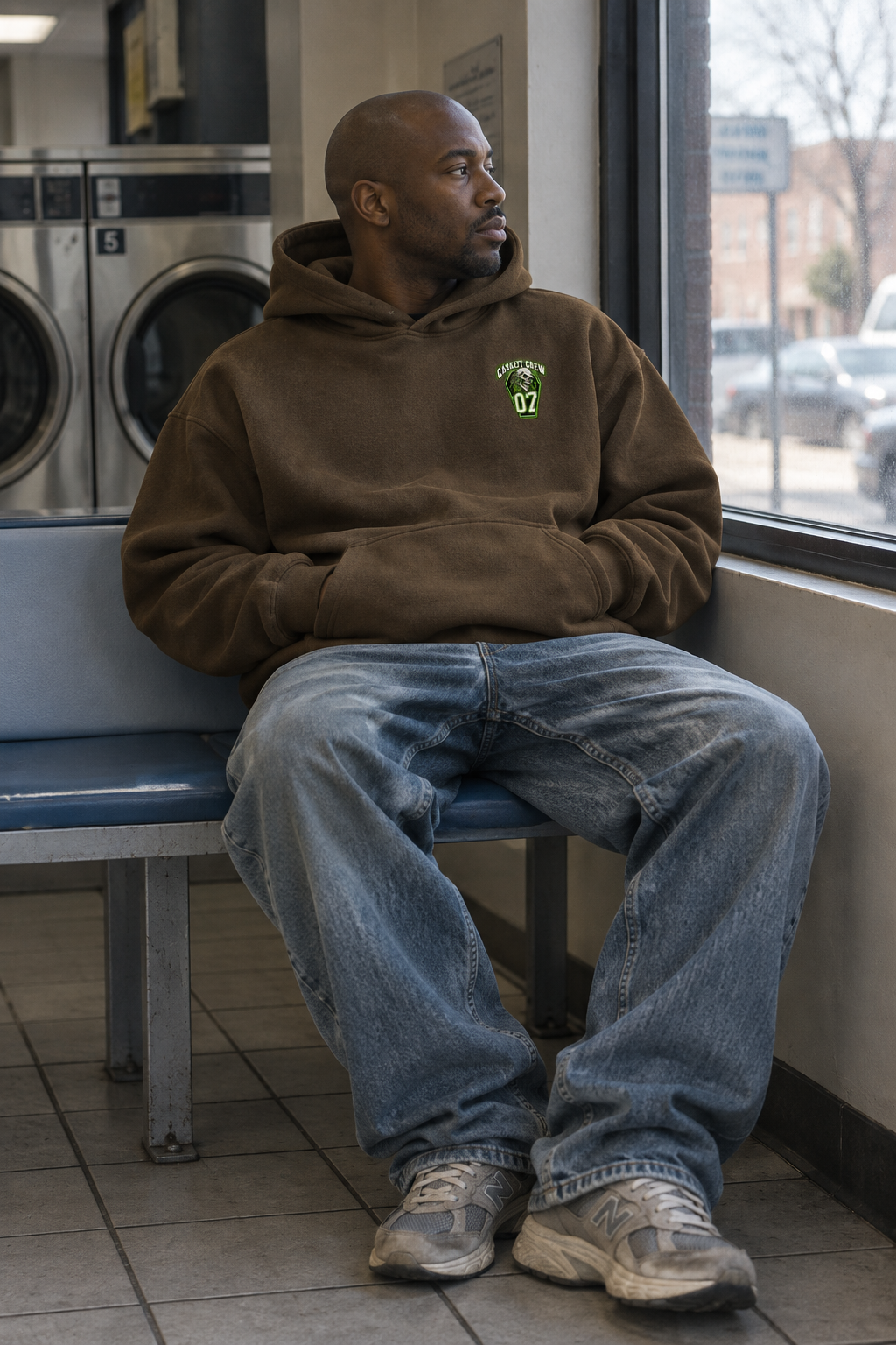 A man sits on a blue bench in a laundromat, wearing an oversized brown hoodie with a small green chest graphic, blue jeans, and gray sneakers.