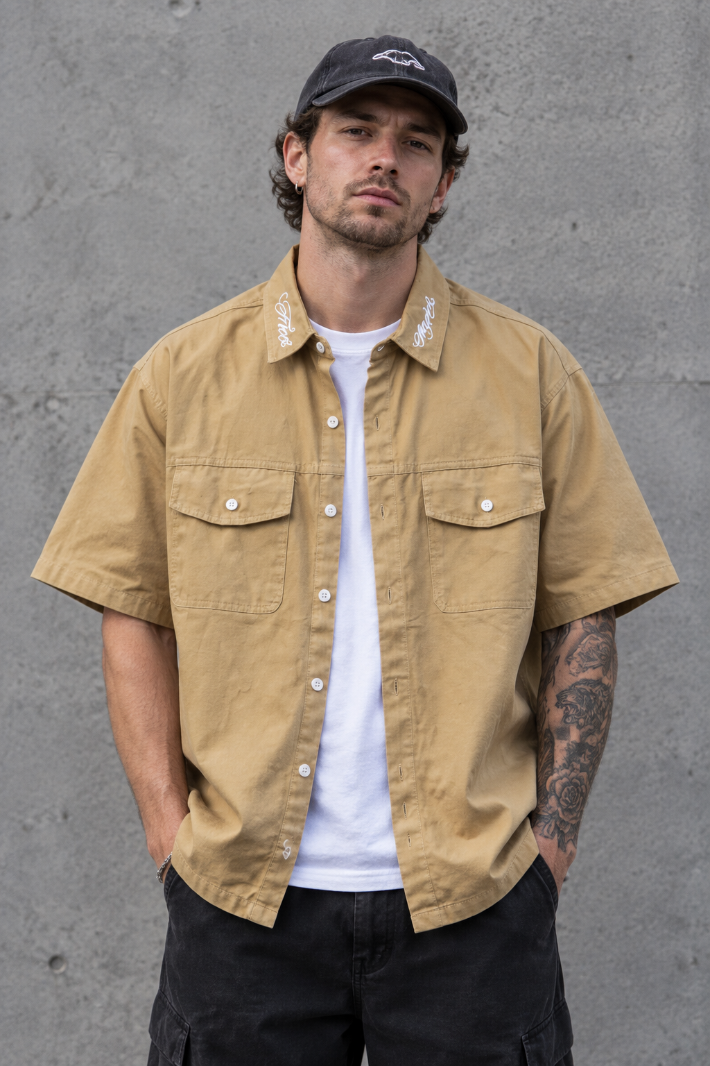 A man wearing a tan short-sleeve button-up shirt with two chest pockets, styled over a white t-shirt and black cargo pants, standing against a textured gray concrete wall.