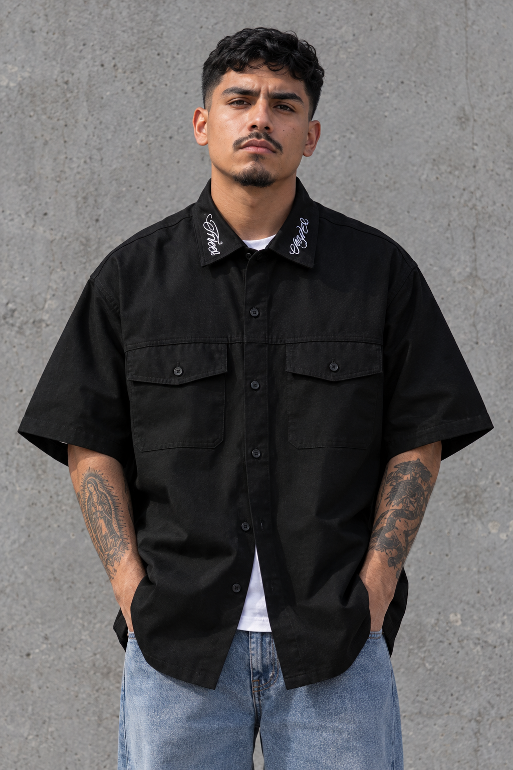 A man wearing a black short-sleeve button-up shirt with two chest pockets and white embroidered details on the collar, paired with light blue jeans, standing against a textured gray concrete wall.