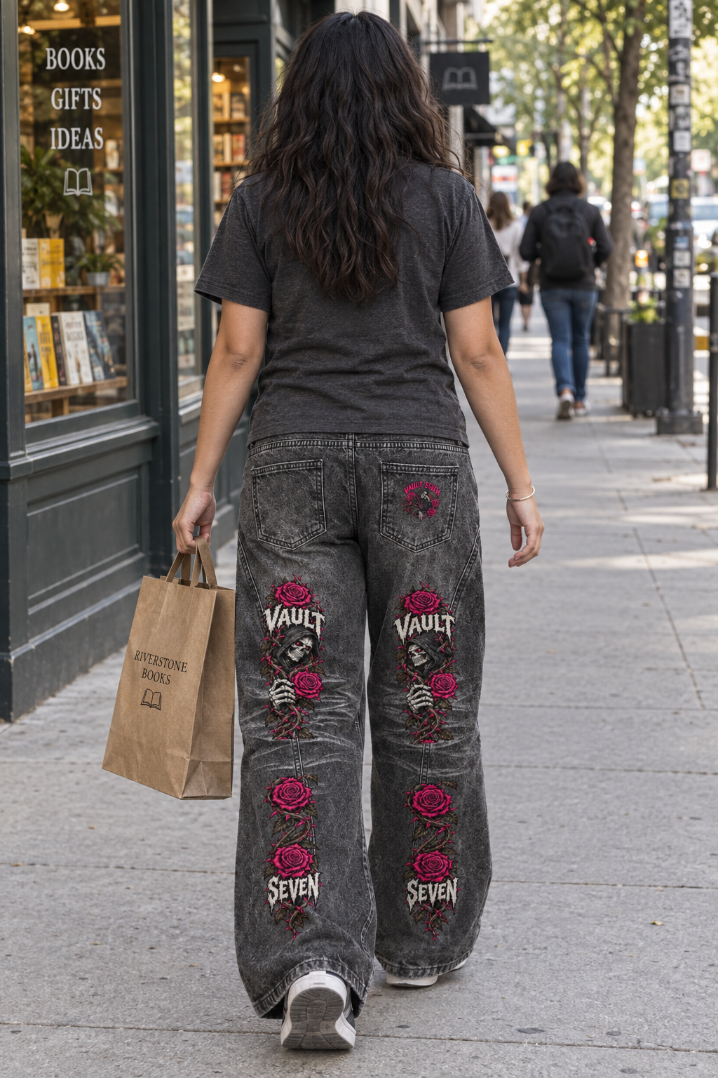 A woman viewed from behind walking on a city sidewalk, wearing faded black wide-leg jeans with large embroidered skull-and-rosette graphics and the word 'VAULT' on the back pockets, along with a dark gray t-shirt and carrying a brown paper shopping bag.