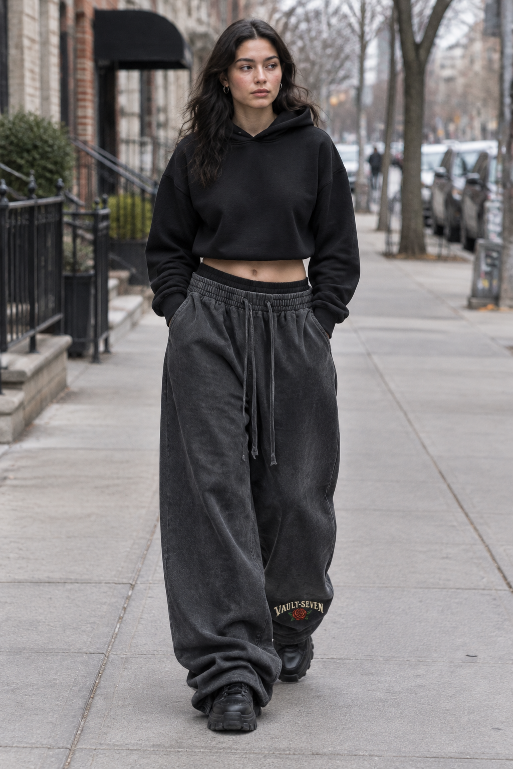 A woman walking on a city sidewalk wearing a black cropped hoodie and dark charcoal gray baggy sweatpants with 'BENET GUTIER' embroidered near the hem.