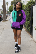A woman stands on a city sidewalk wearing a color-block hoodie with purple, black, mint green, and light gray panels, paired with black-and-white sneakers and white socks.