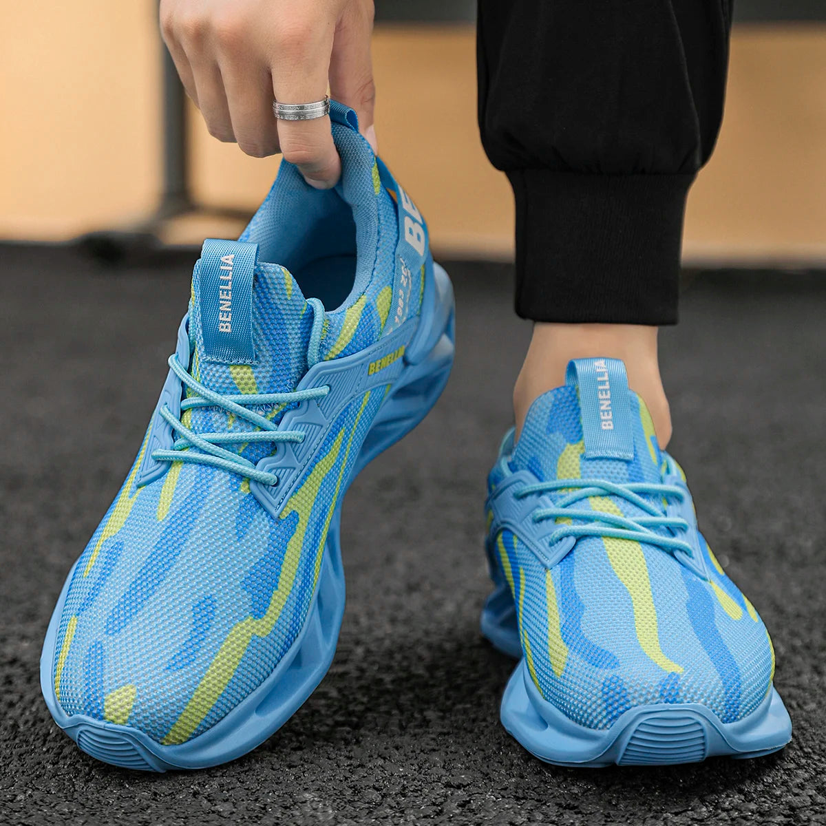 A person wearing light blue mesh sneakers with yellow abstract stripes and light blue laces, with one hand lifting the heel of the left shoe. The shoes have 'BENELLIA' branding on the tongue.