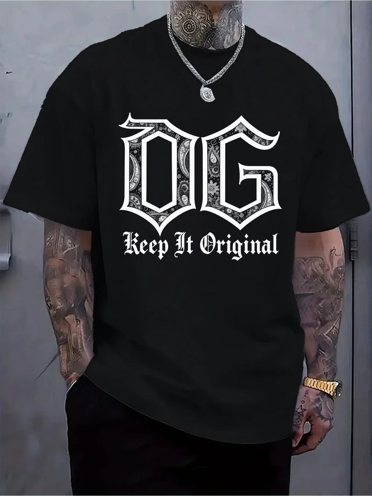 A man wearing a black short-sleeve t-shirt with a white gothic-style 'DG' graphic and the words 'Keep It Original' printed on the front. The man has visible tattoos on his arms and neck, wearing a silver chain necklace and a gold watch.