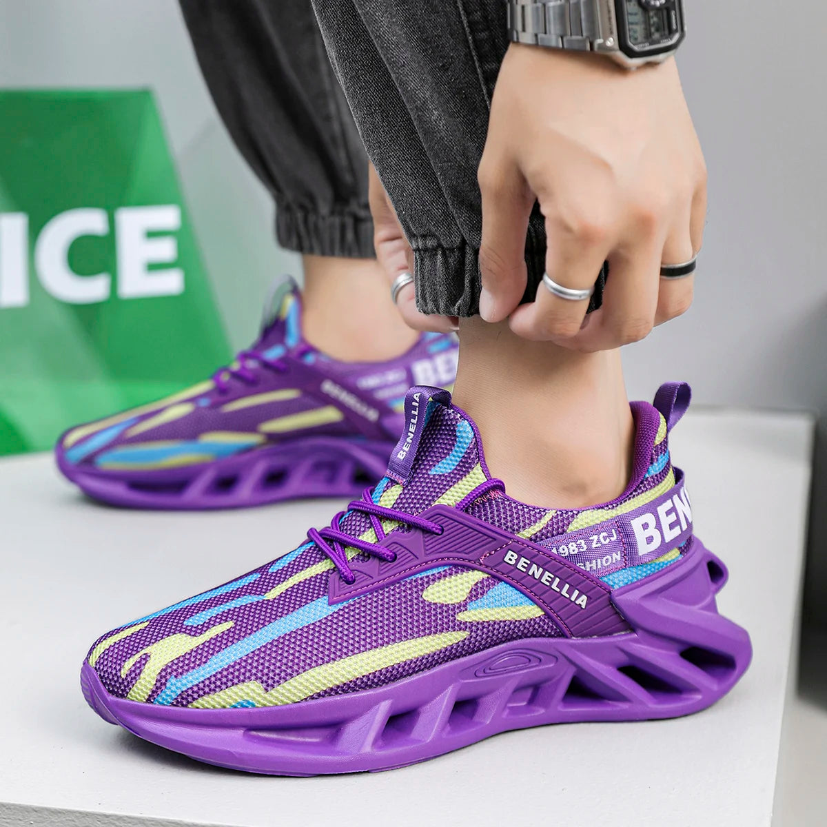 A person wearing purple athletic sneakers with multicolor accents, shown from the waist down on a light gray surface. The shoes feature a purple mesh upper with yellow and light blue graphic stripes, a purple chunky sole, and 'BENELLIA' branding.