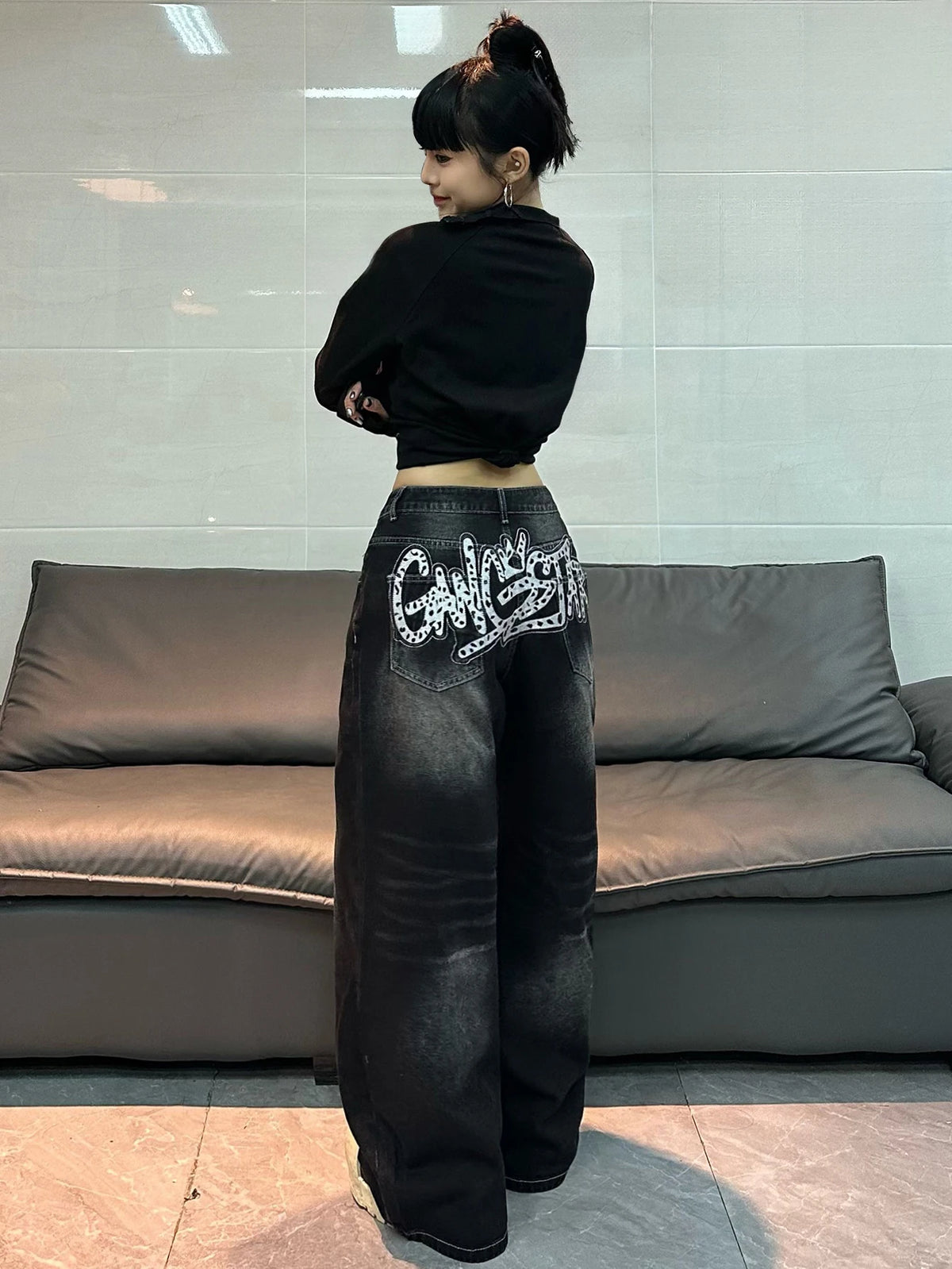 A person standing in front of a gray leather sofa, wearing a black cropped long-sleeve top and very wide-leg black jeans with a large white graffiti-style graphic on the back pocket.