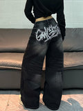 A person shown from the back wearing wide-leg black denim jeans with a large white graphic text design on the back pocket, paired with a black cropped long-sleeve top, standing in front of a gray couch.