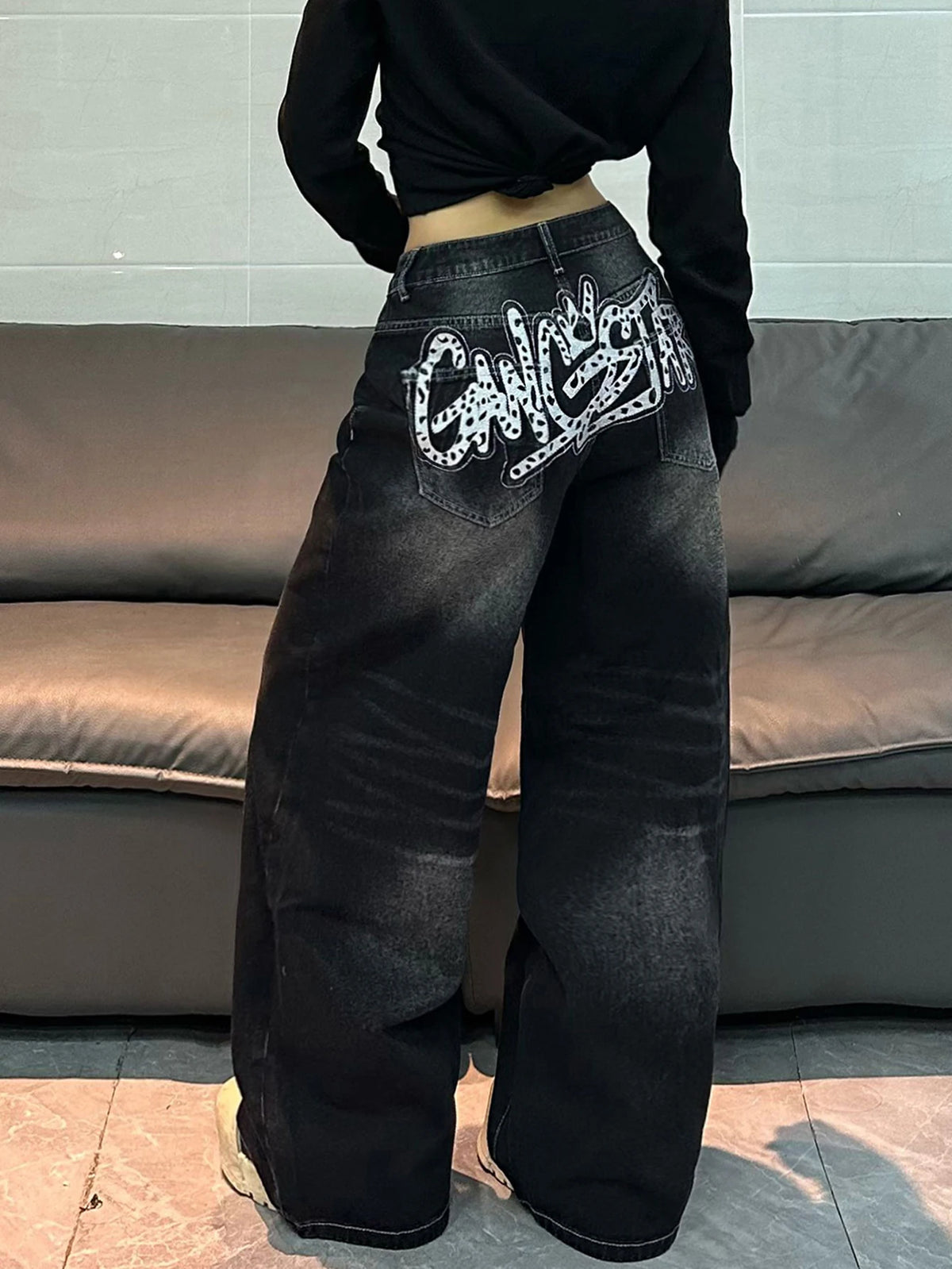 A person shown from the back wearing wide-leg black denim jeans with a large white graphic text design on the back pocket, paired with a black cropped long-sleeve top, standing in front of a gray couch.