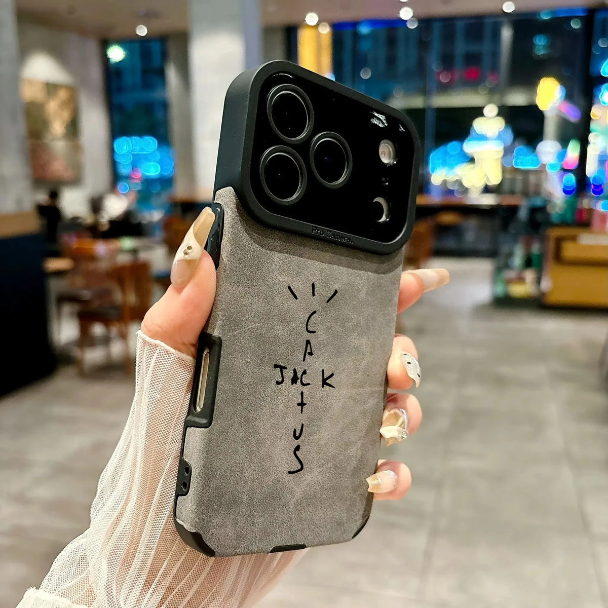 A hand holding a gray textured phone case with a black camera bumper and black side edges, featuring the handwritten text 'I CAN JACK US' on the back. The background is a blurred indoor cafe environment with warm lighting and bokeh.