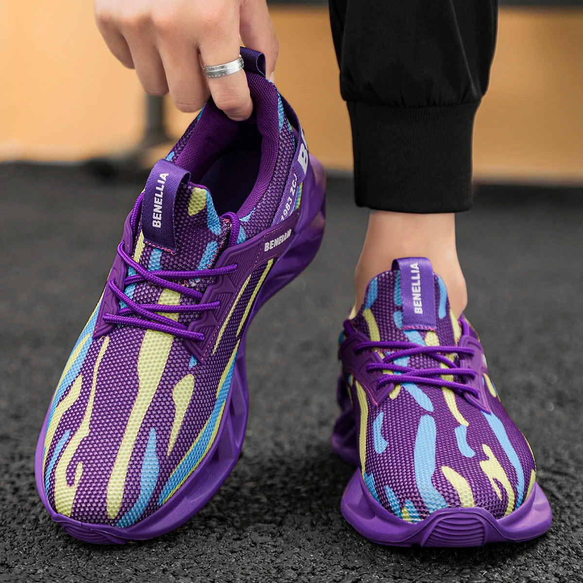 Purple mesh athletic sneakers with a wavy abstract pattern in light blue and pale yellow, shown on a person standing on a dark textured floor while one hand lifts the heel of the left shoe. The brand name BENGHELLA is visible on the tongue.