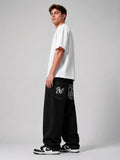 A model wearing a white t-shirt, black denim jeans with 'BW' embroidery and patterned pocket details, and black-and-white sneakers, standing in profile against a light gray studio background.