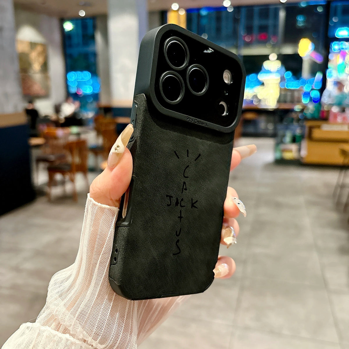 A hand holding a black textured phone case with a custom hand-drawn style logo that reads 'Cactus Jack' in black marker, set against a blurred cafe background.