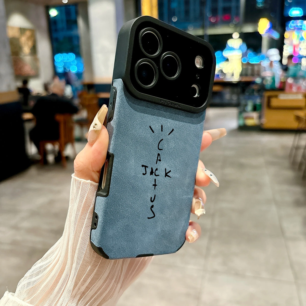 A hand holding a light blue suede-textured phone case with a black camera bumper and black handwritten text reading 'I CAN JACK US' in a blurred cafe setting.