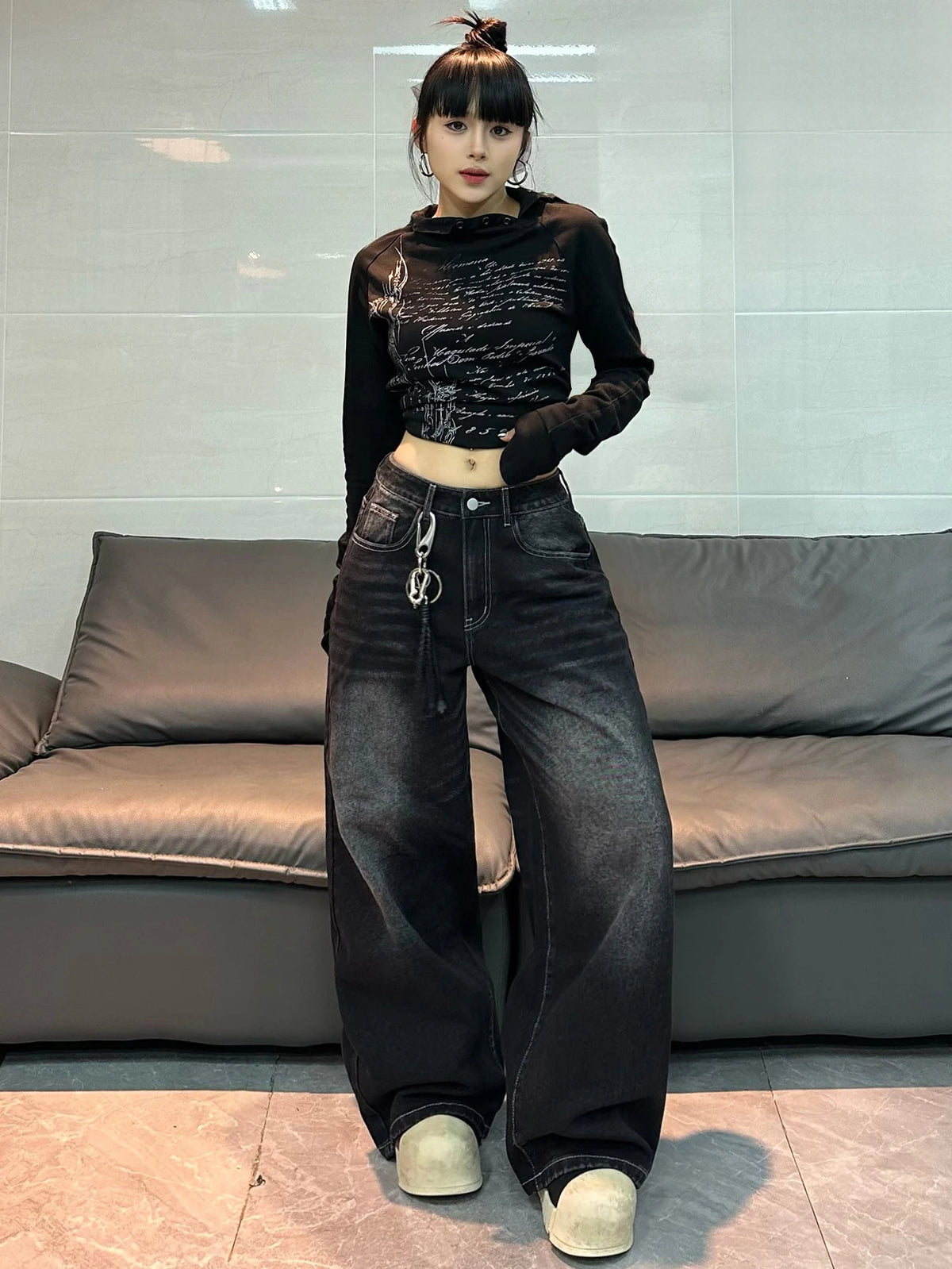 A person stands indoors in front of a gray sofa wearing a black long-sleeve cropped top with a graphic print and very wide-leg dark jeans with a drawstring waist and silver chain detail.