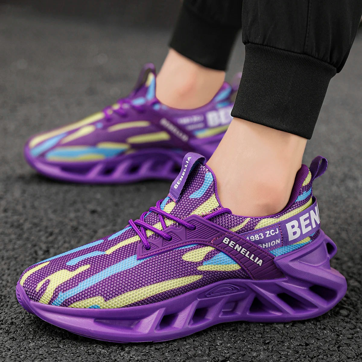 A close-up of a person wearing purple mesh sneakers with yellow and light blue abstract stripes, standing on a dark asphalt surface. The shoes feature a chunky sculpted purple sole and the brand name 'BENELLIA' printed on the side.
