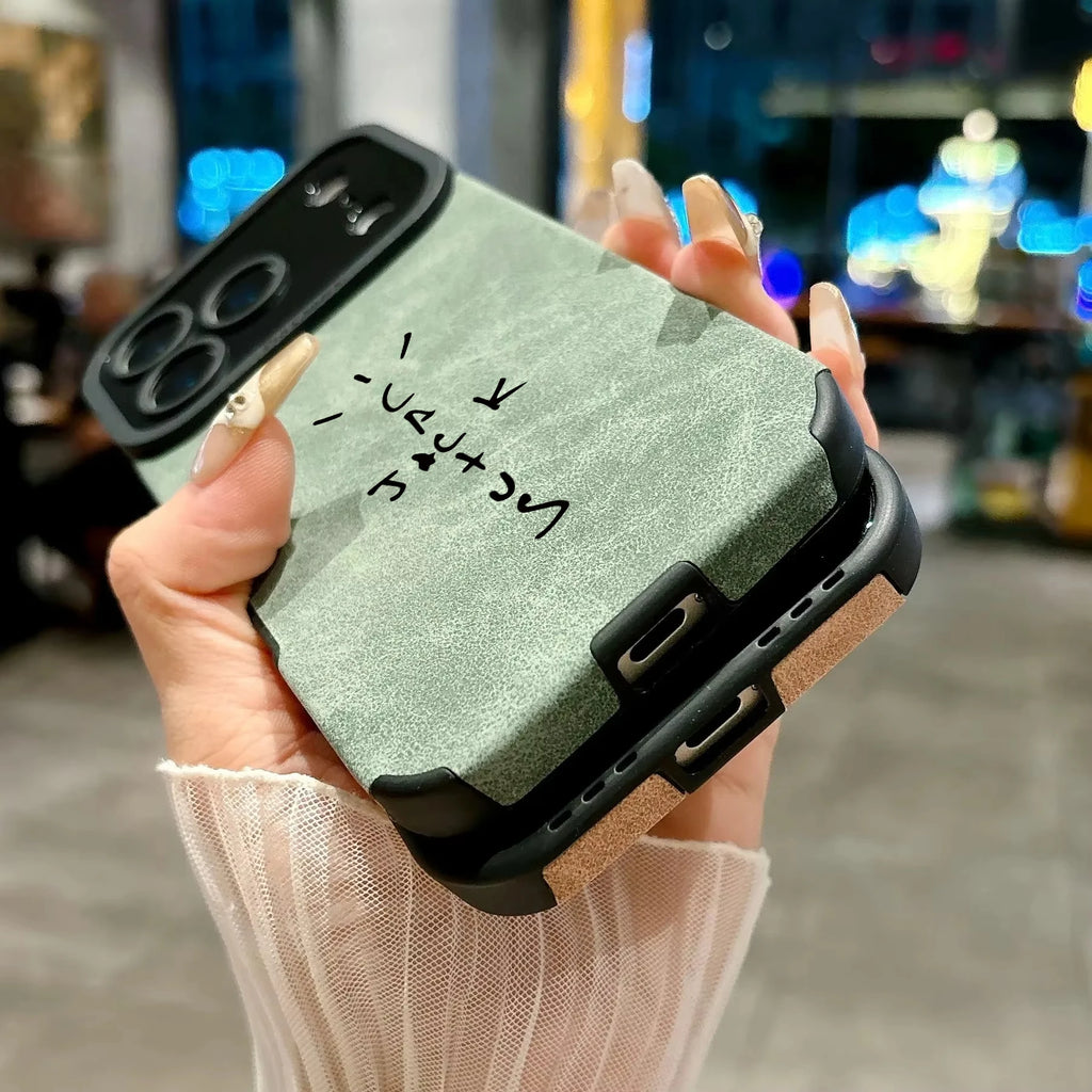 A close-up of a hand holding a rugged phone case with a sage green textured back, black protective corners and camera surround, and a tan side bumper. The hand has long manicured nails, and the background is a blurred indoor setting with warm lighting.