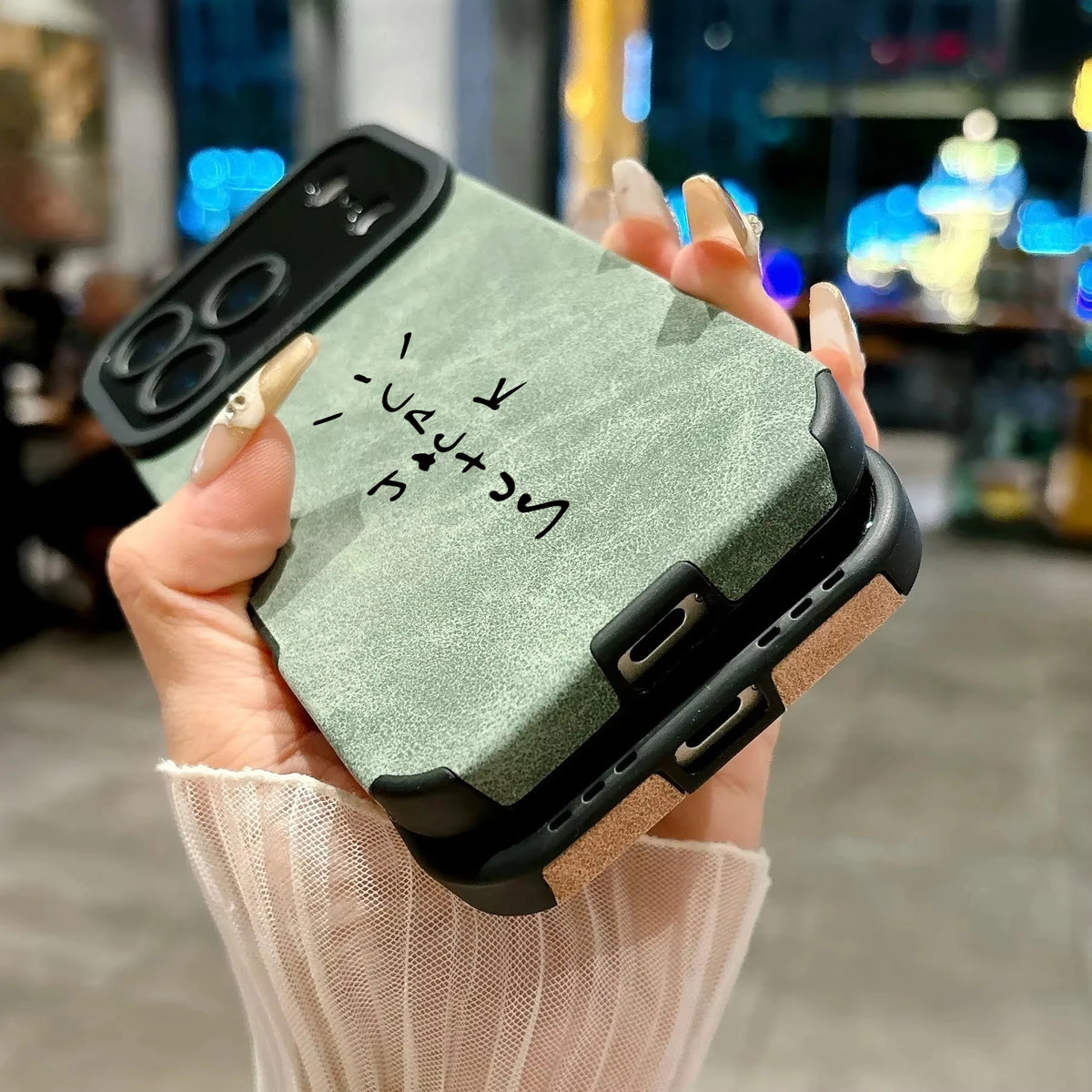 A close-up of a hand holding a rugged phone case with a sage green textured back, black protective corners and camera surround, and a tan side bumper. The hand has long manicured nails, and the background is a blurred indoor setting with warm lighting.
