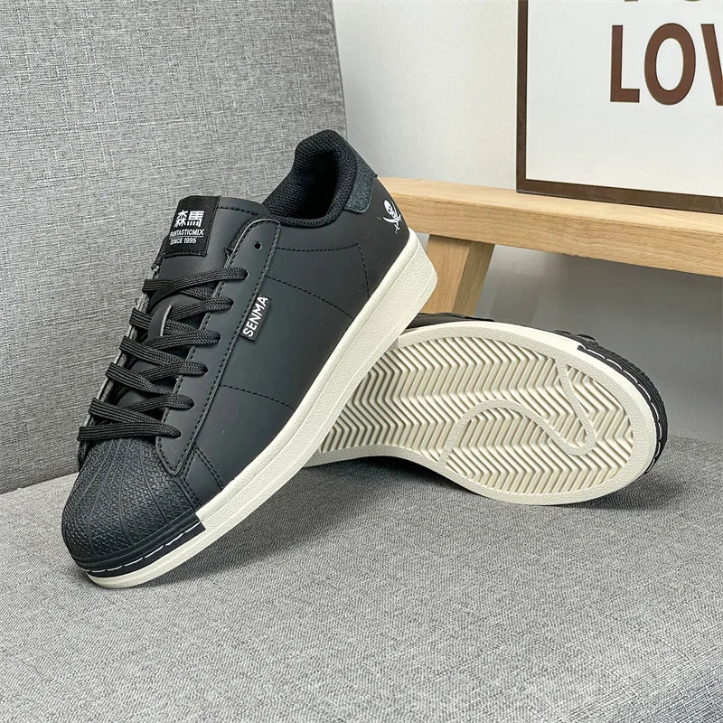 A pair of black leather sneakers with white rubber soles, one standing upright and the other lying on its side to show the sole tread. The shoes are placed on a gray textured fabric surface next to a wooden shelf.