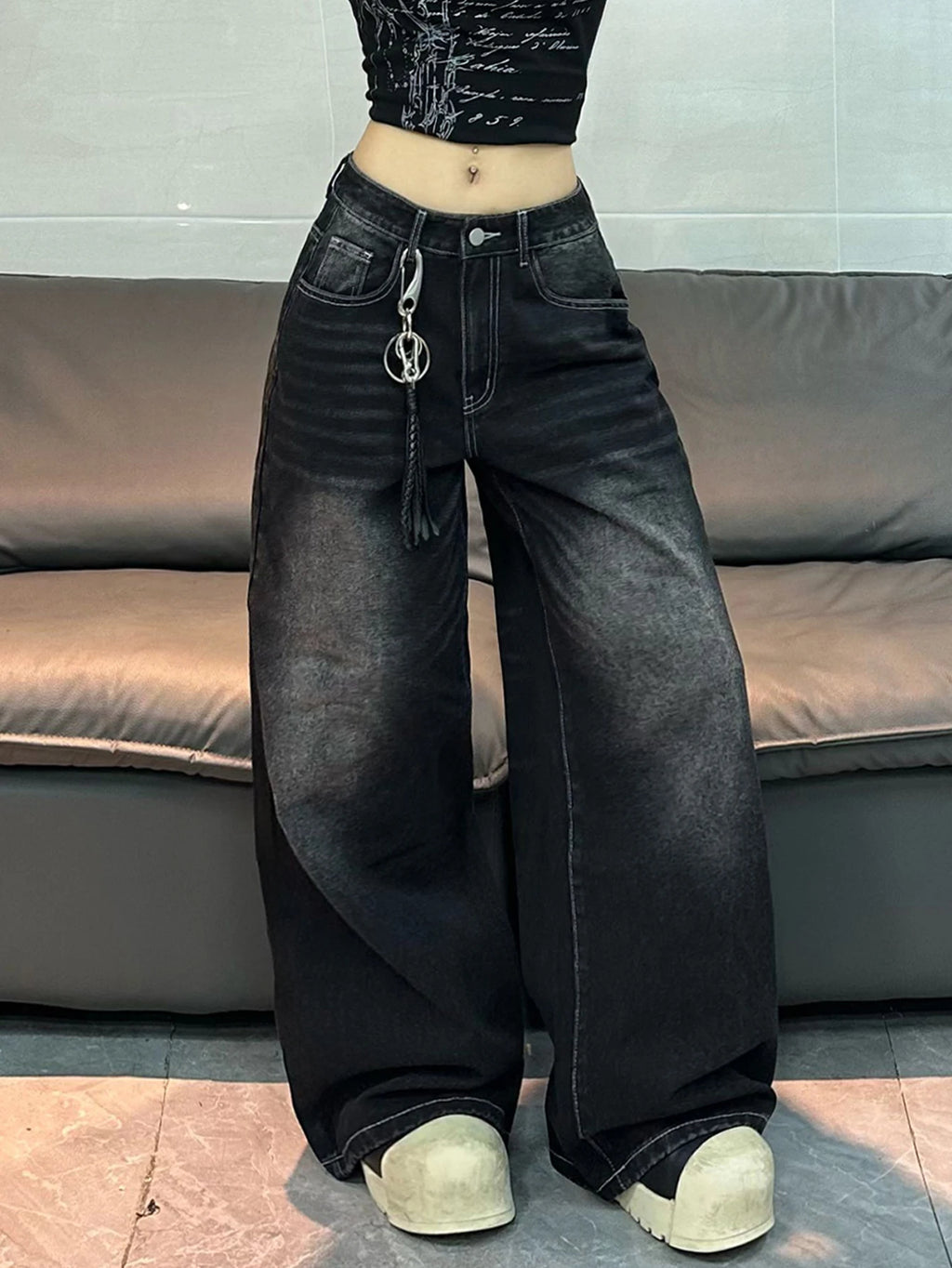 A person wearing very wide-leg black denim jeans with a faded gray wash, styled with a black cropped top and beige sneakers, standing in front of a gray leather sofa.