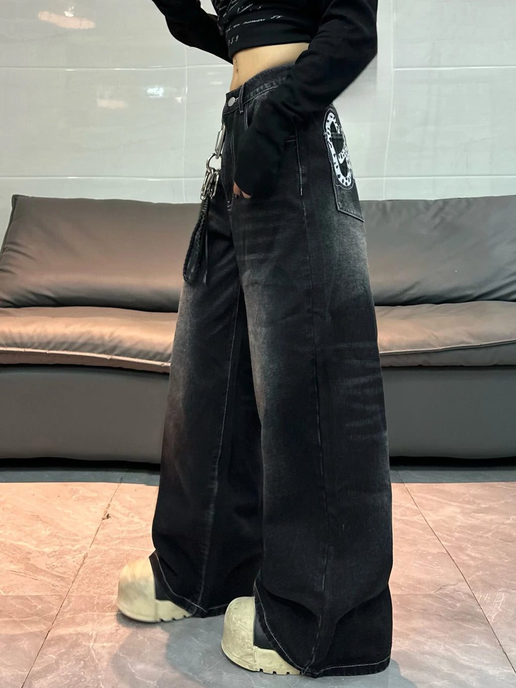 A person wearing wide-leg black washed denim jeans with a circular graphic patch on the back pocket, a black cropped long-sleeve top, and chunky off-white platform shoes, standing in front of a gray sofa.