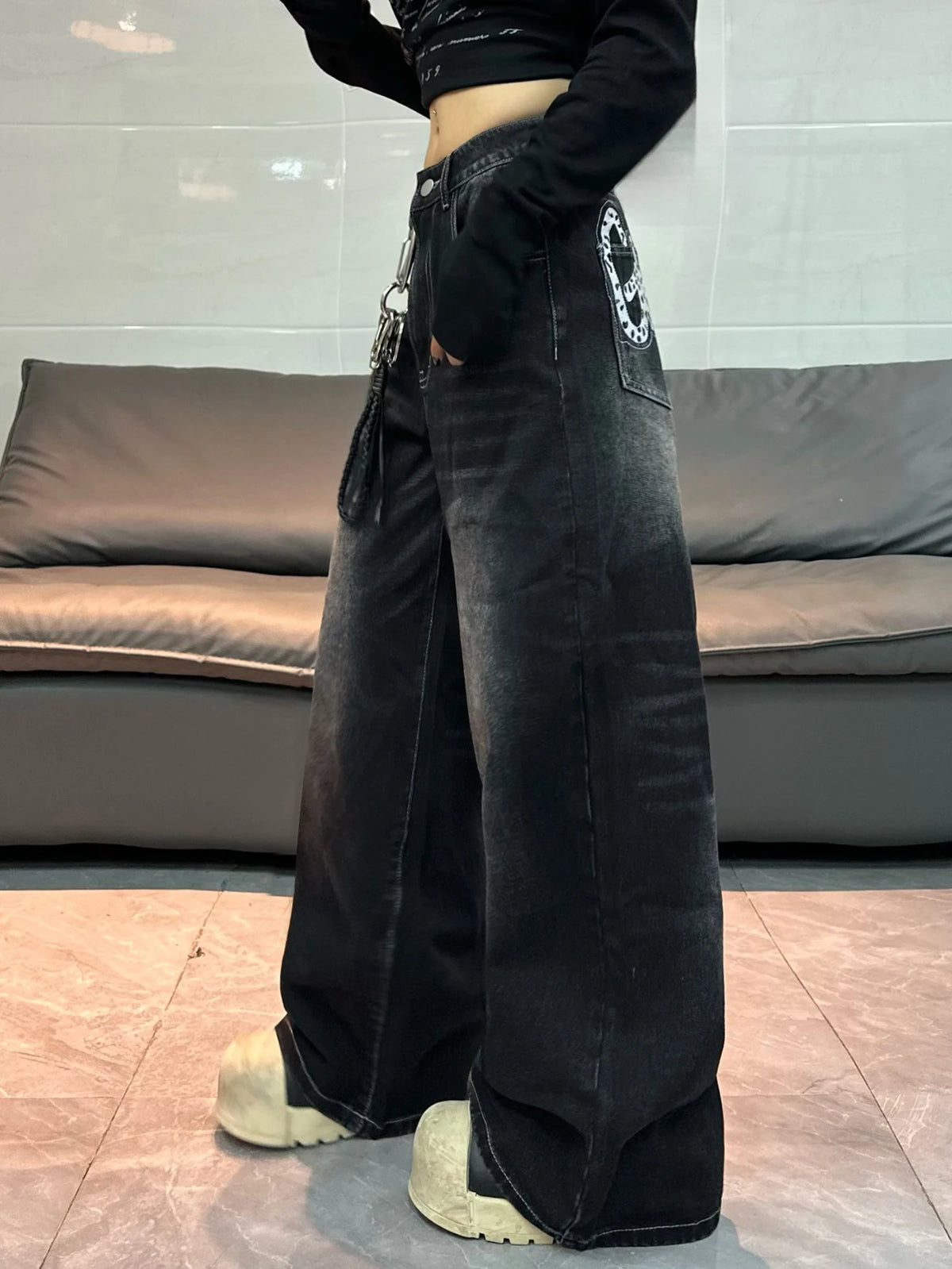 A person wearing wide-leg black washed denim jeans with a circular graphic patch on the back pocket, a black cropped long-sleeve top, and chunky off-white platform shoes, standing in front of a gray sofa.
