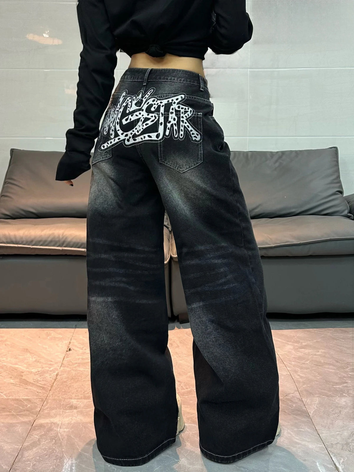 A person wearing wide-leg black denim jeans with a large white graphic print on the back pocket, standing in front of a gray sofa.