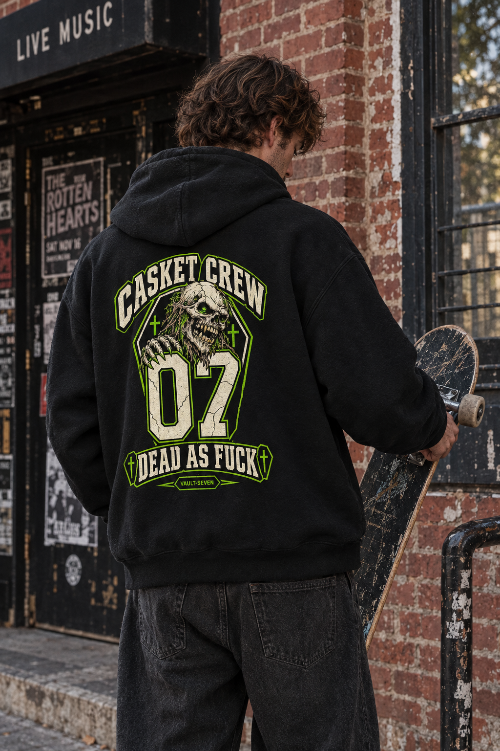 A person standing outdoors in front of a brick wall and storefront, wearing a black hoodie with a large green and white graphic on the back that reads 'CASKET CREW 07 DEAD AS FUCK' with a skull illustration. The person is holding a skateboard.