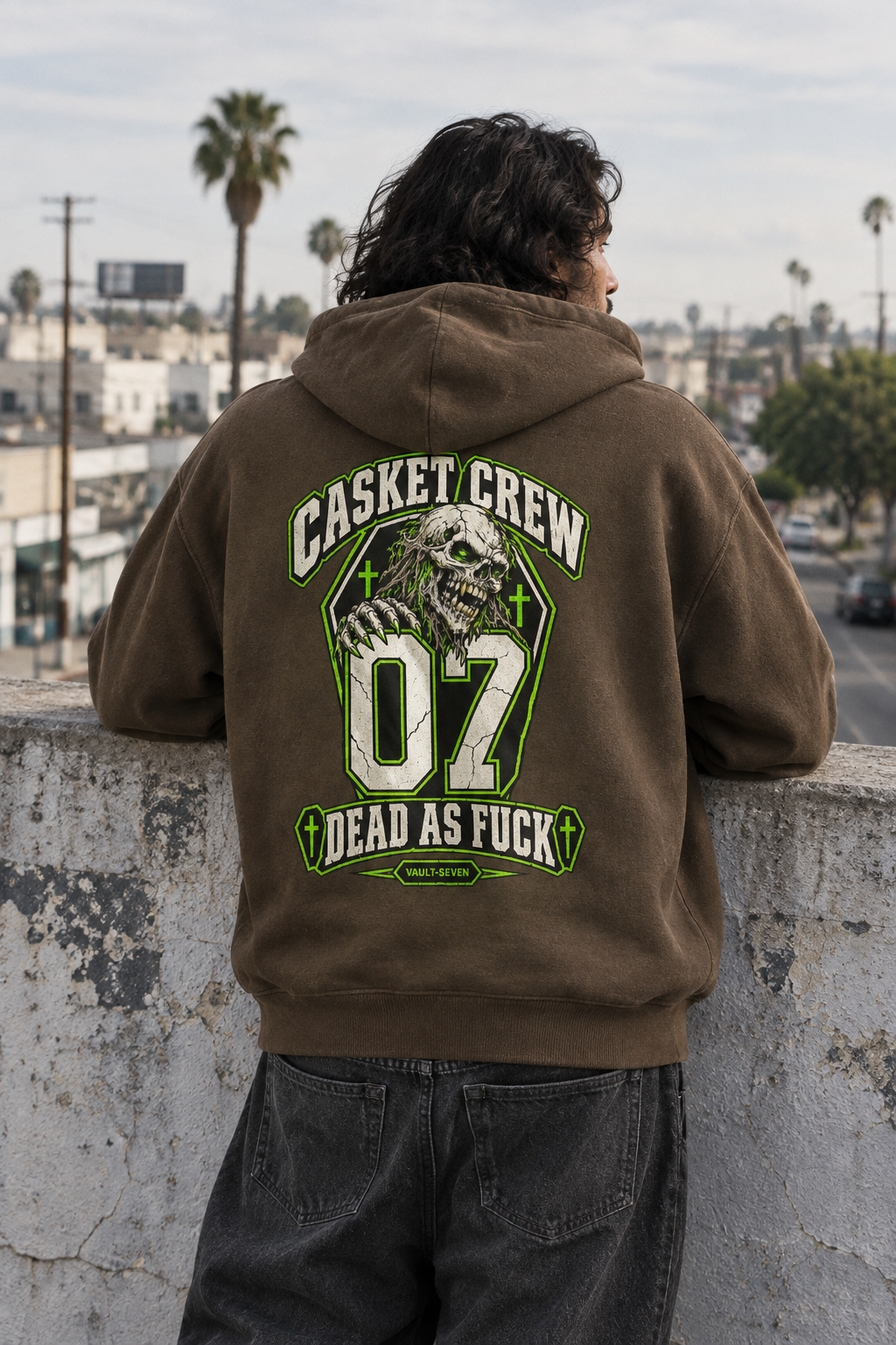 A person viewed from behind wearing a brown hoodie with a large green and white graphic on the back that reads 'CASKET CREW 07 DEAD AS FUCK' with a skull illustration, standing outdoors on a concrete ledge overlooking a city street.