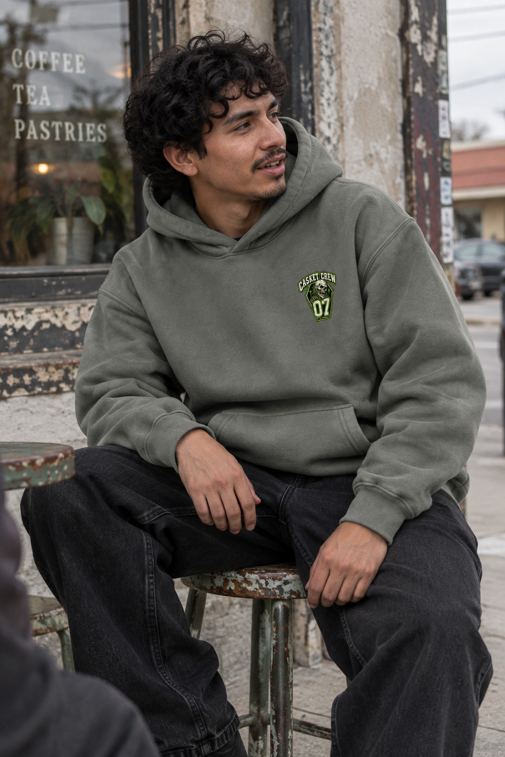 A man wearing a muted olive green hoodie with a green and yellow embroidered graphic on the left chest, sitting on a metal stool in front of a rustic storefront.