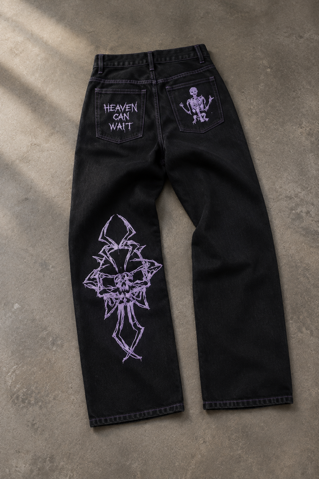 Black denim jeans laid flat on a concrete floor, featuring purple graphic prints including a large skull design on one leg, a skeleton on the other back pocket, and 'HEAVEN CAN WAIT' text on the third pocket.