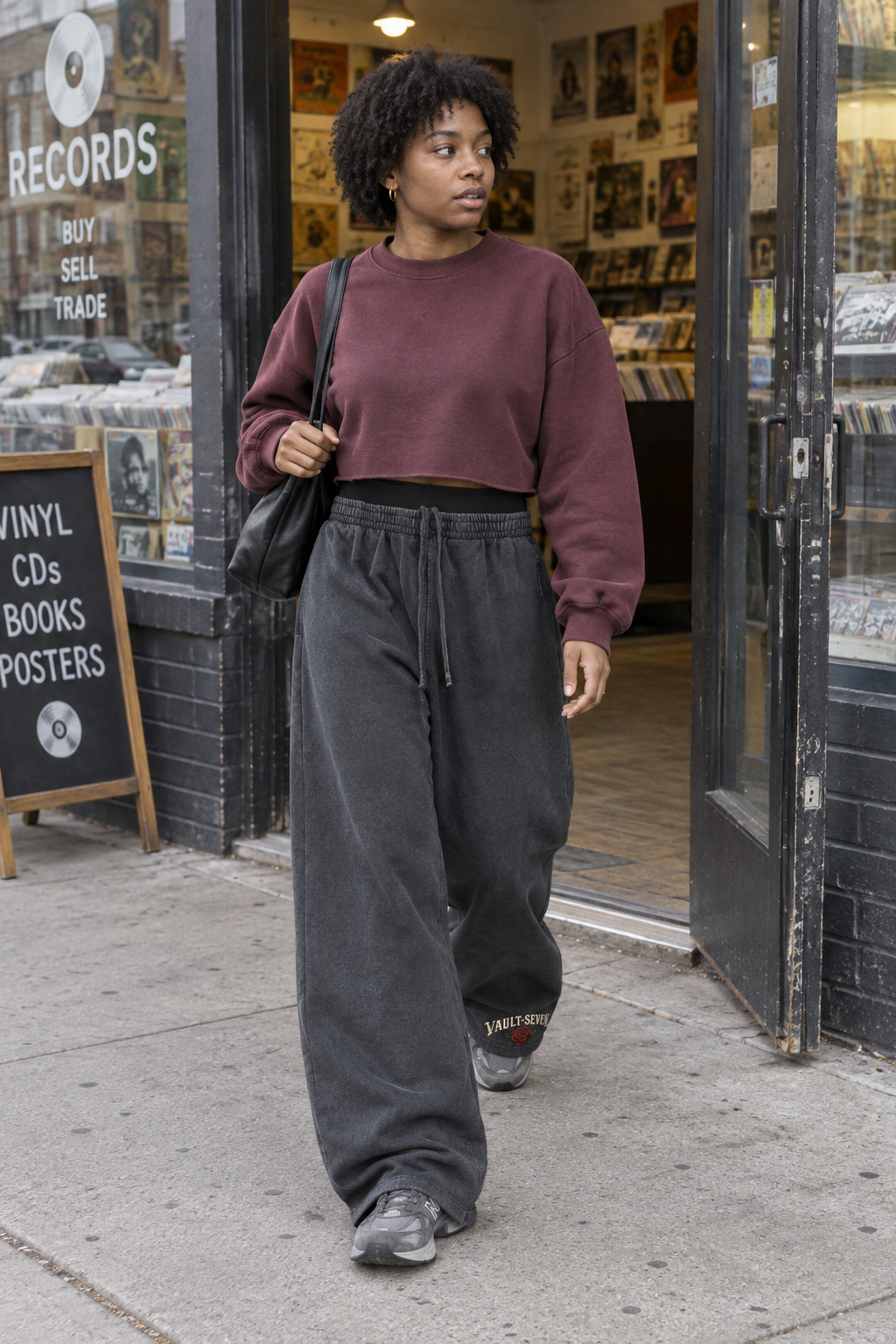 A woman walking out of a record store on a city sidewalk, wearing a maroon cropped crewneck sweatshirt and loose dark charcoal drawstring sweatpants.