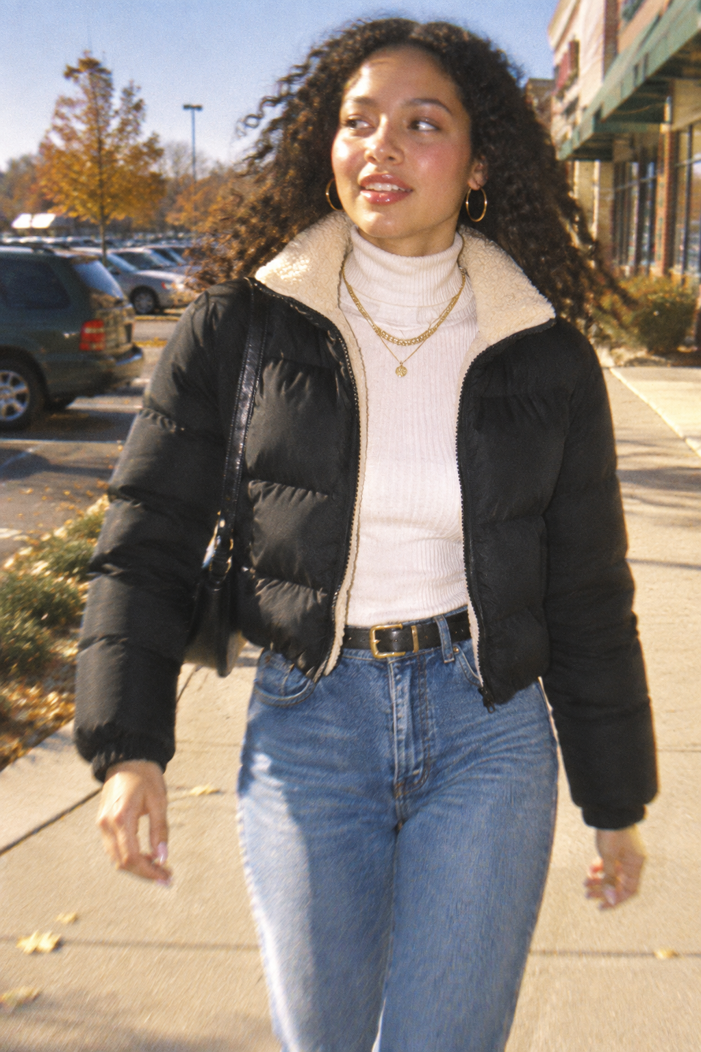 A woman walking outdoors on a sidewalk wearing a black puffer jacket with a cream sherpa collar, a white ribbed turtleneck, blue jeans, and gold jewelry.