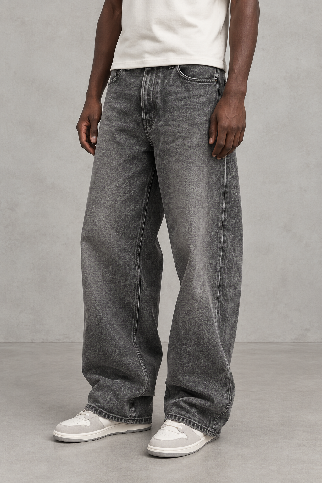 A person wearing loose-fit gray denim jeans with a faded wash, paired with a white t-shirt and white sneakers, standing against a textured gray studio wall.