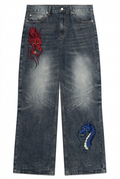 A pair of dark blue acid-wash jeans with embroidered dragon motifs on the left thigh in red and on the right thigh in blue and white.