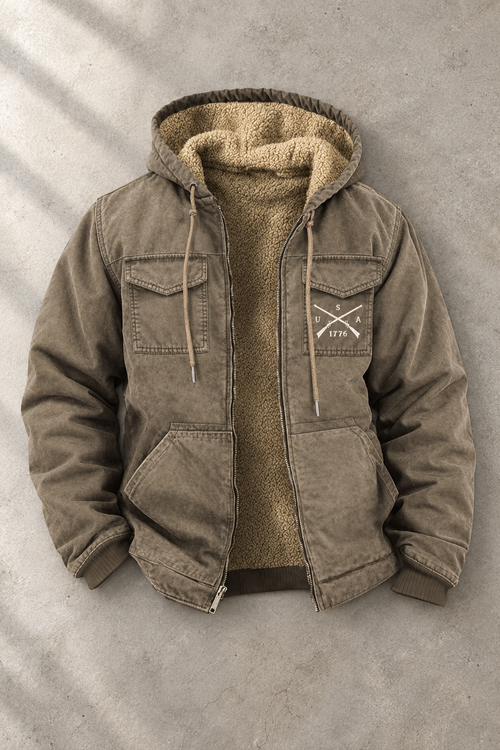 An olive brown hooded jacket with a tan sherpa lining, laid flat on a textured gray concrete background. The jacket features a zippered front, drawstring hood, and a small white embroidered logo on the left chest.