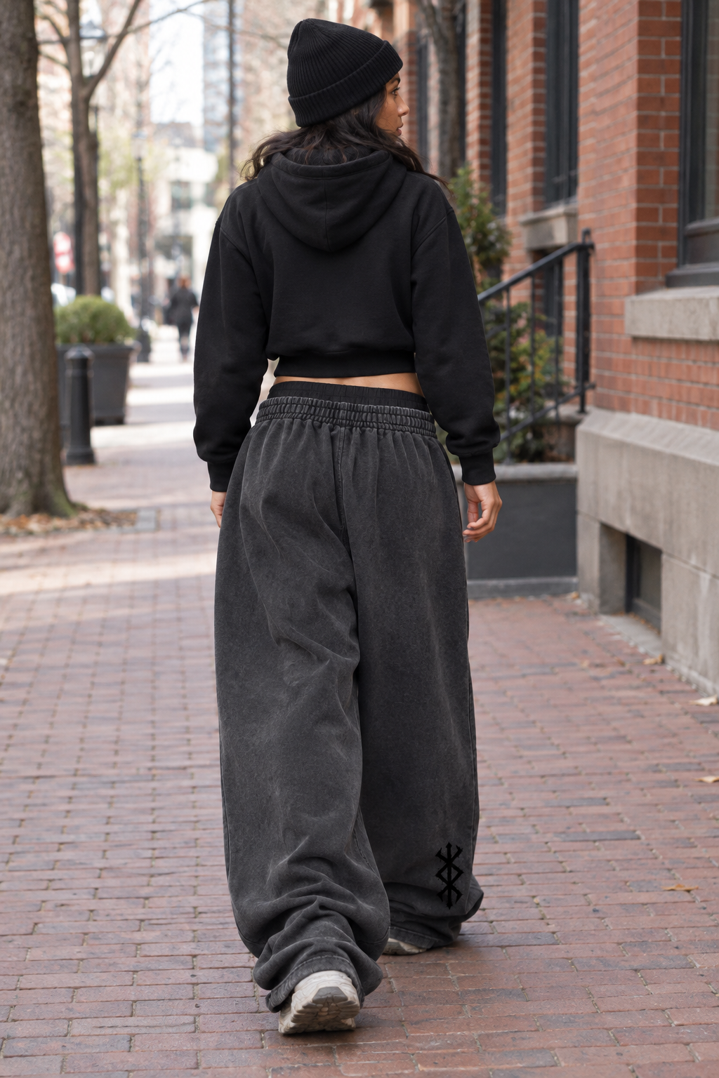 A person walking away on a brick sidewalk, wearing a black cropped hoodie, black beanie, and very wide-leg dark gray sweatpants with a small black graphic near the hem.